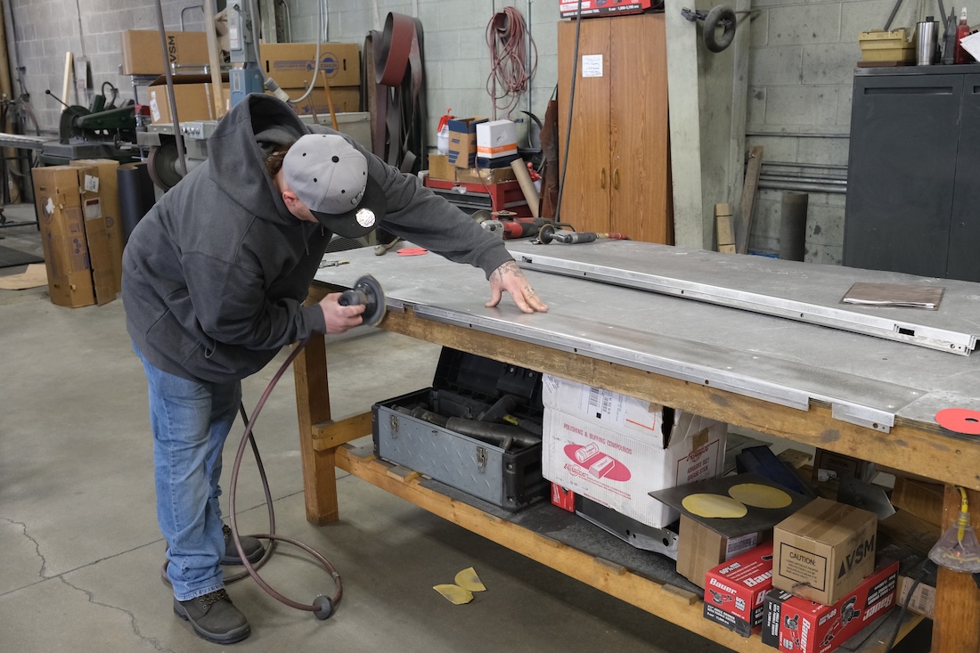 A person in a workshop uses a handheld power sander on a long metal panel placed on a wooden workbench, surrounded by tools, boxes, and industrial equipment.