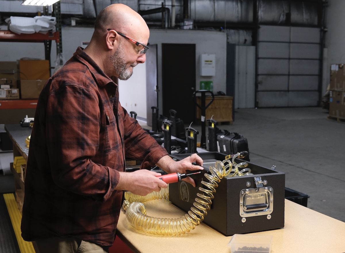 A person wearing safety glasses uses a handheld power tool to work on a black equipment unit with a coiled yellow hose in an industrial workshop.
