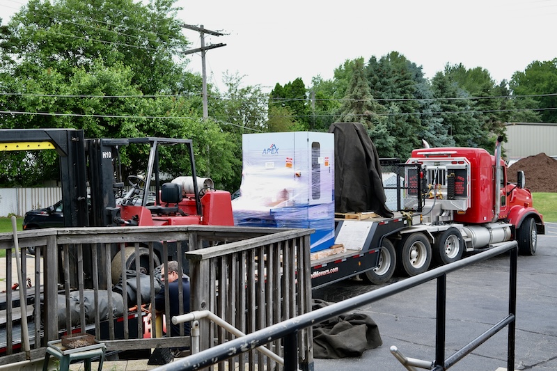 A red forklift beside a red semi-truck with a flatbed trailer carrying a shrink-wrapped APEX machine, while workers secure it in a parking lot.