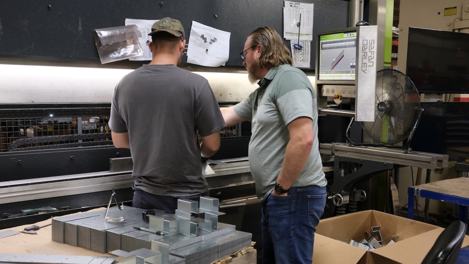 Two workers discuss a metal bending machine beside stacked metal parts in a fabrication workshop.