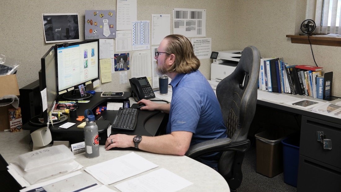 Jimmy Hansen works at a computer in an office with papers, binders, and printed schedules around his desk.