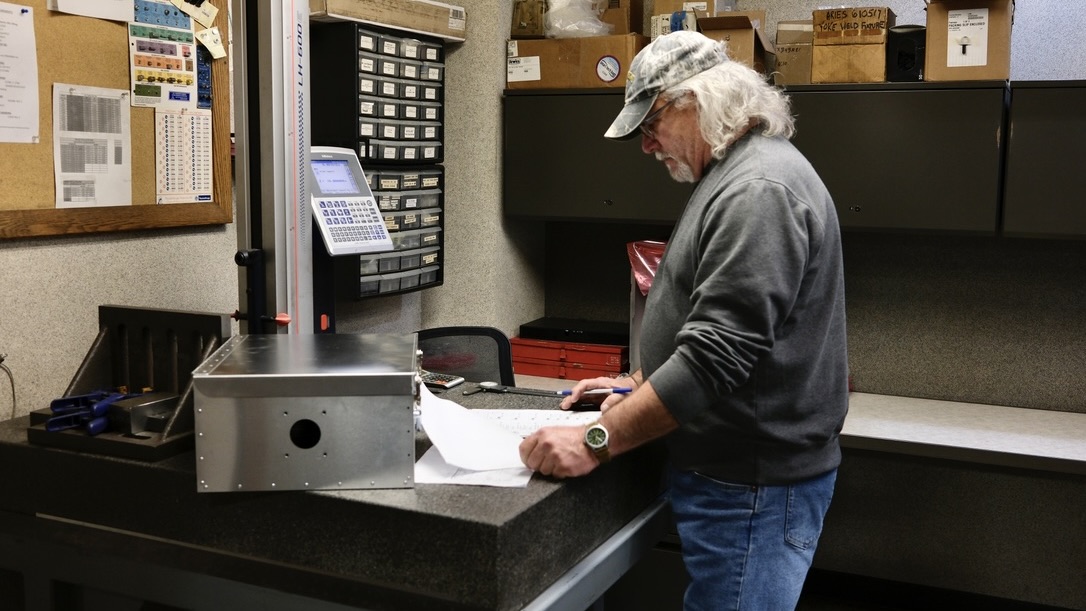 A man reviews paperwork beside a measuring machine and metal enclosure in a workshop inspection area.