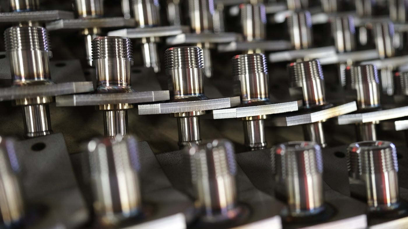 Rows of polished threaded metal fittings attached to flat brackets are arranged in a manufacturing workspace.