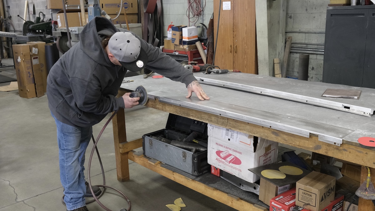 A worker uses a handheld grinder to smooth the edge of a large metal panel on a workshop table.