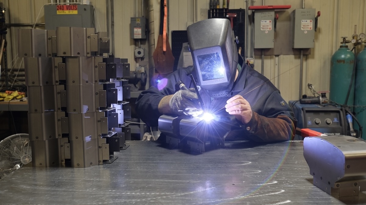 A welder wearing a protective helmet works on a metal component, creating a bright welding arc on a workshop table.