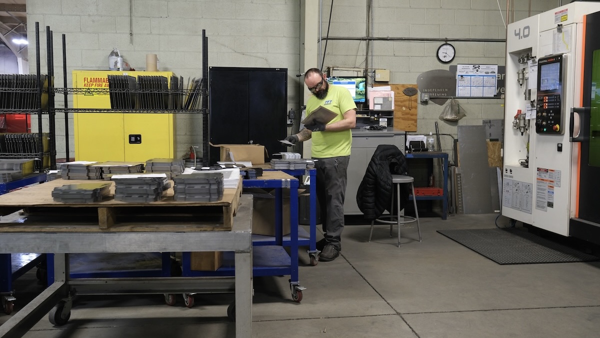 A worker in safety glasses inspects stacked metal parts beside a large industrial machine in a manufacturing shop.