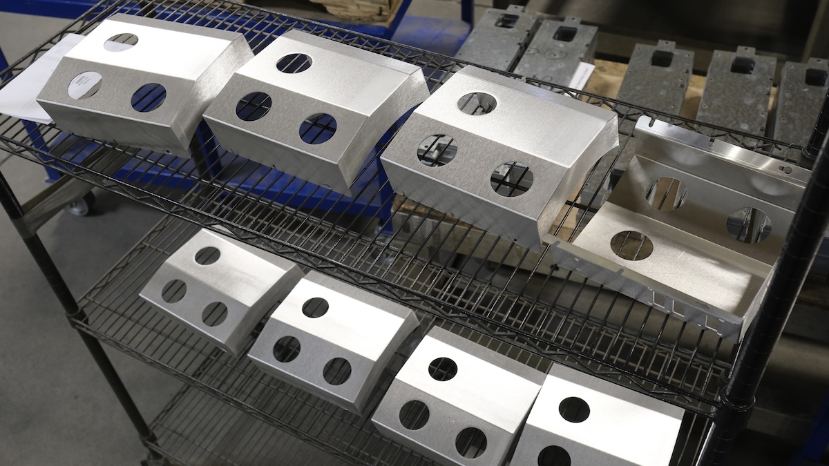 Brushed metal enclosures with circular cutouts are arranged on wire shelving in a manufacturing shop.