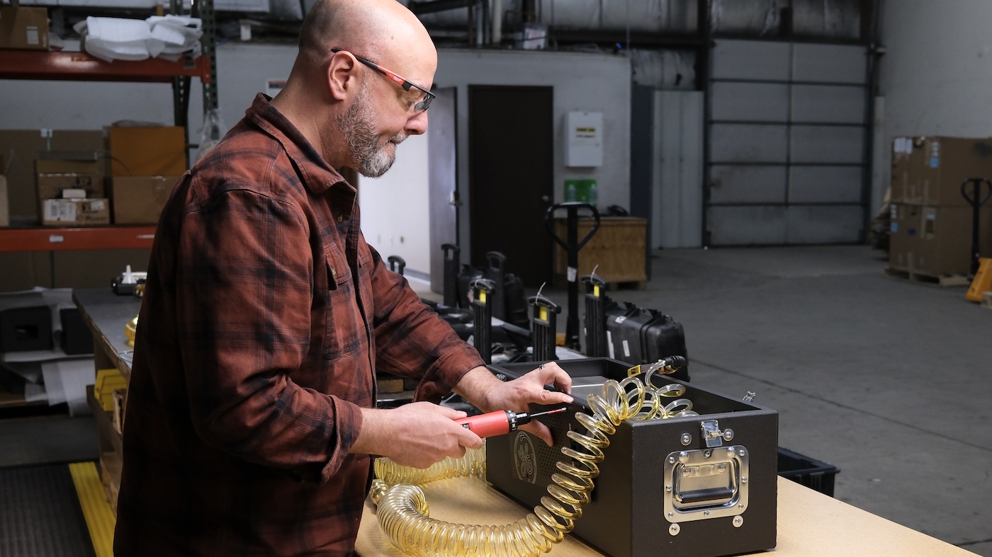 A man wearing safety glasses uses a screwdriver to assemble a black equipment case with a coiled yellow hose attached.