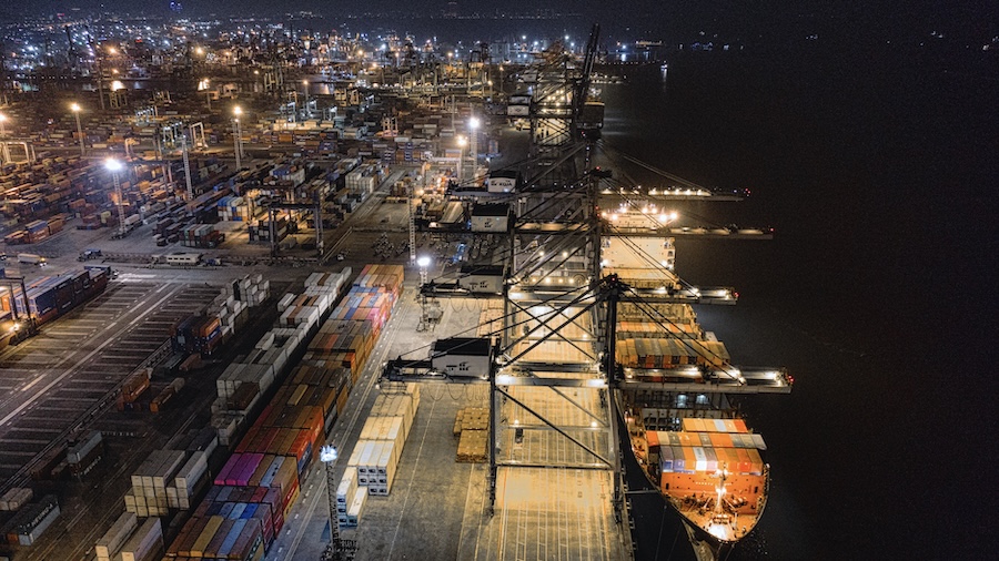 A brightly lit container port at night with cargo cranes, stacked shipping containers, and a ship docked along the water.